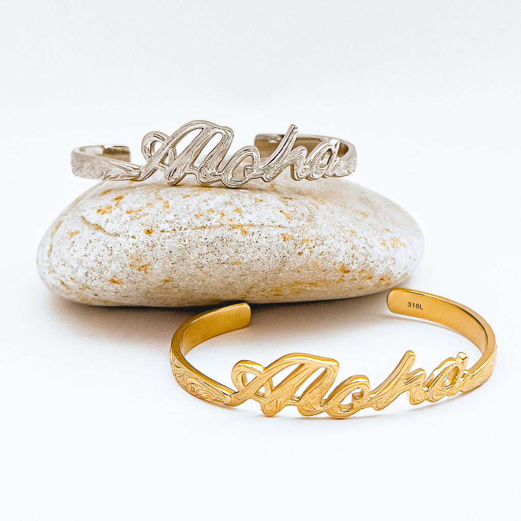 Two bangle bracelets shown, one gold-plated bangle and one stainless steel bangle (color silver). Cuff style bracelet carved script Aloha with engraved design motif