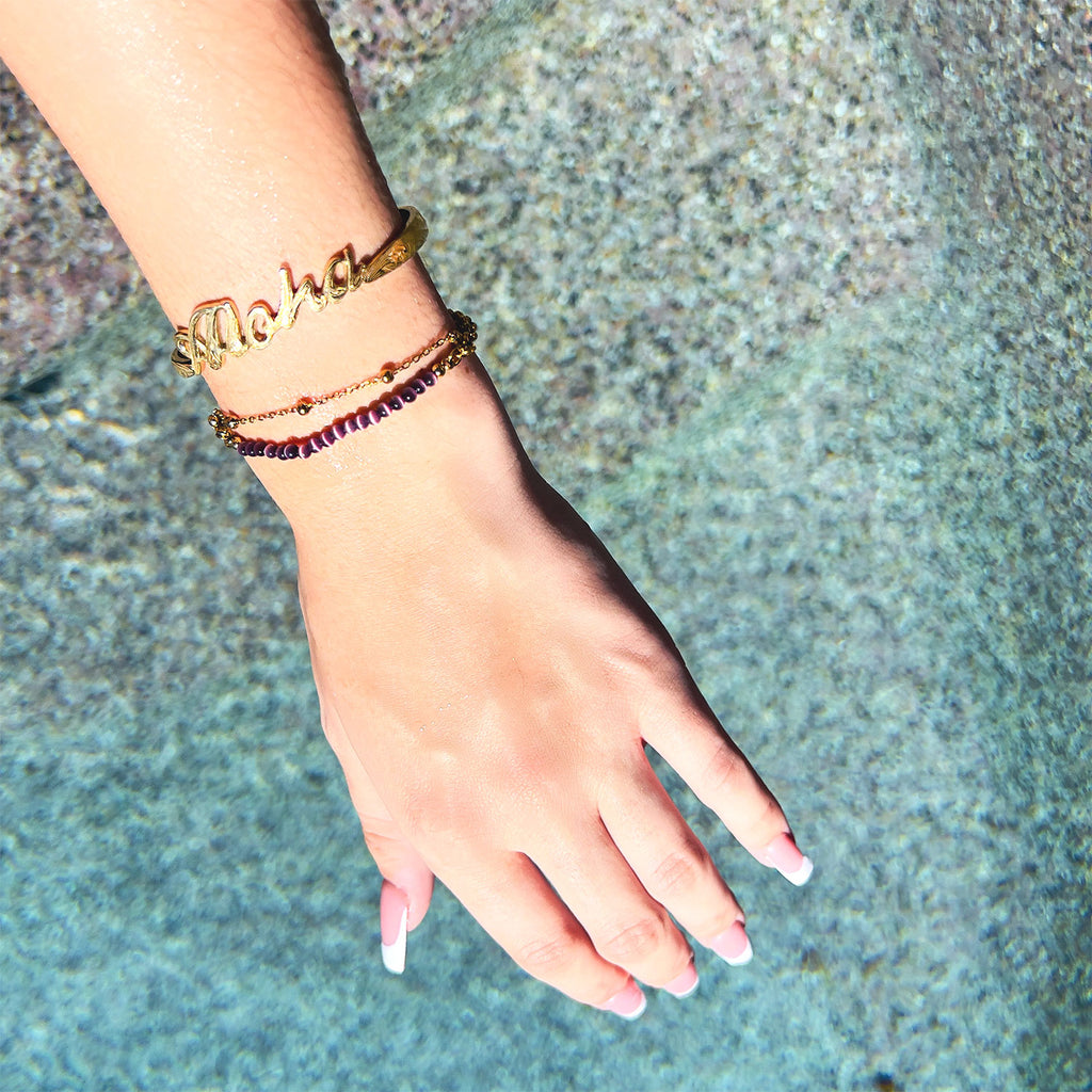 Girls hand over water with gold-plated "Aloha" carved bracelet. Layered with purple beaded bracelet.