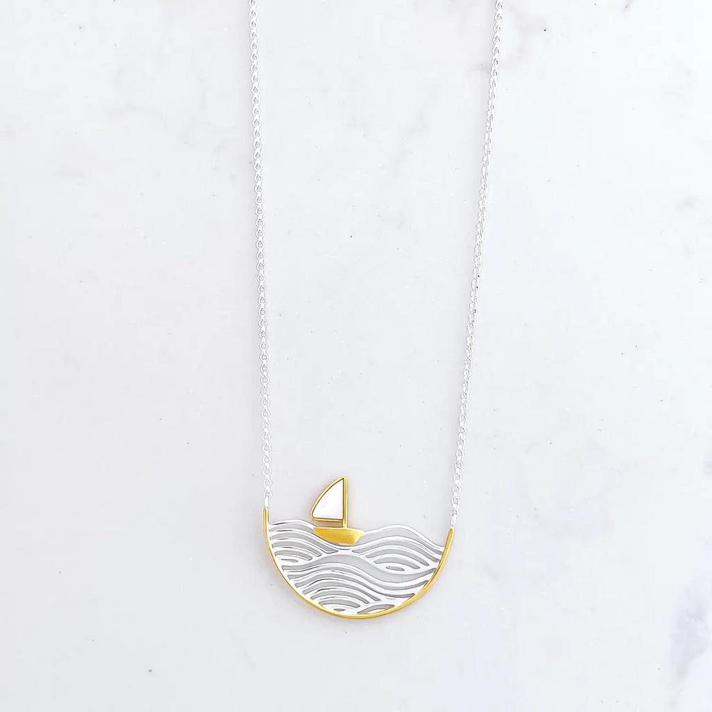 Sterling silver two-tone (silver and gold) sailboat pendant necklace, silver chain. Pendant is a half circle. In the pendant is silver engraved waves, sitting on the wave is a small sail boat figure that is gold with a white sail. The rim of the half circle is also gold.