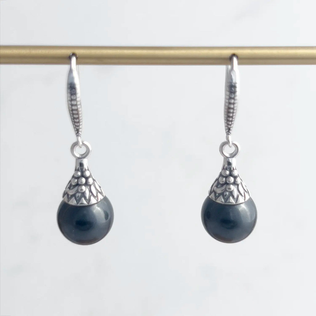 Black freshwater pearl and sterling silver drop earrings with a fishhook backing. Sterling silver has a pattern engraved. Earrings shown on a display.