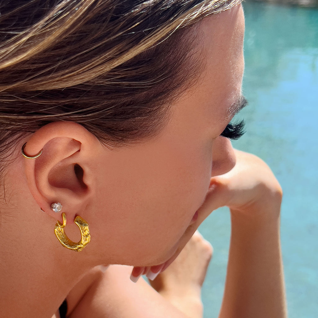 Girl sitting by pool wearing 18k gold plate 'driftwood' textured hoop earrings. Hoop ends 3/4 way back up to top.