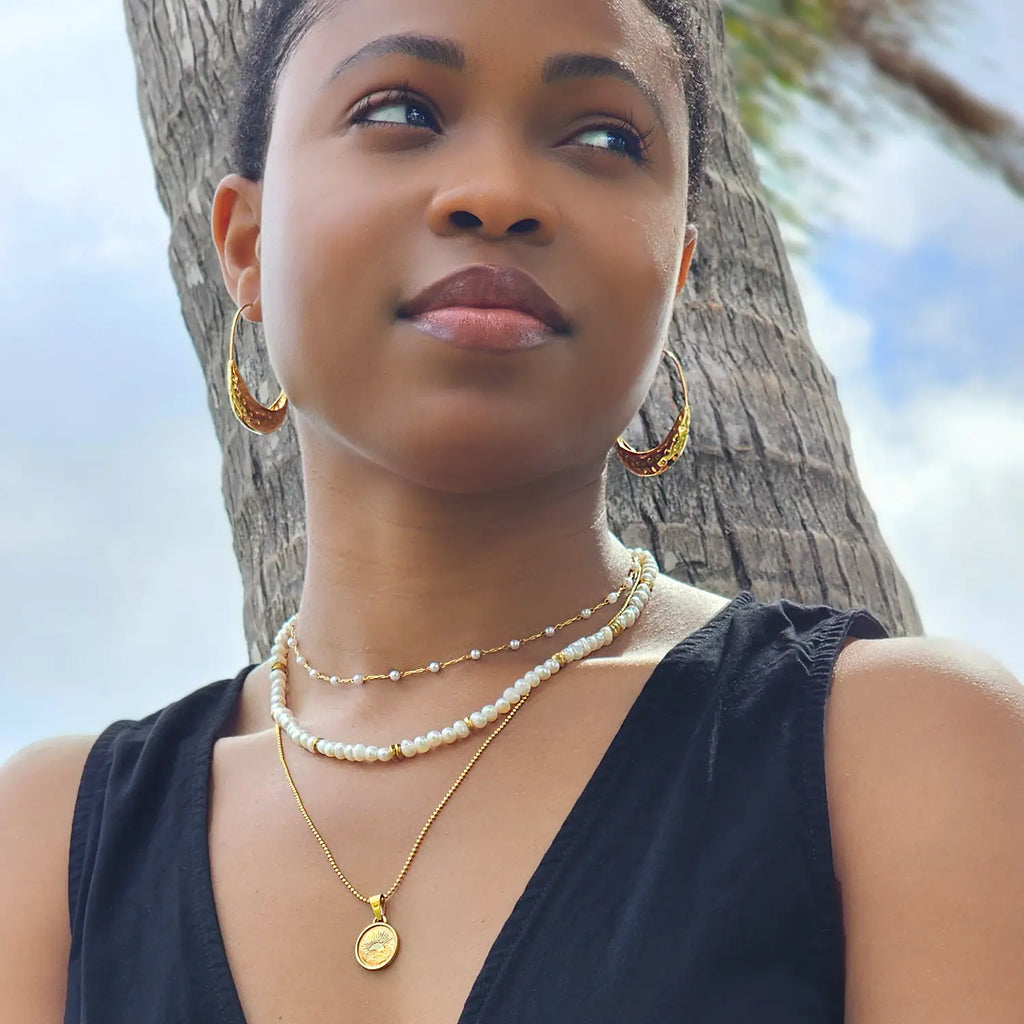 Girl standing in front of palm tree wearing freshwater pearl beaded necklace with 18k gold plated beads spaced out a few times along the necklace. Necklace layered with longer gold pendant chain and smaller pearl beaded choker necklace.