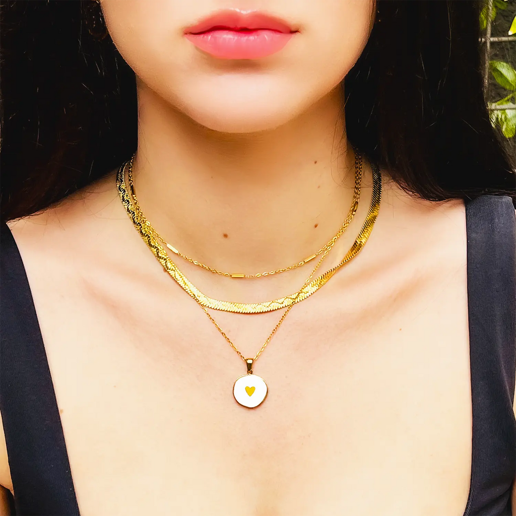 18k gold plated pendant necklace, white enamel center and small gold heart. Shown on a girl wearing black tank, layered with gold snake chain and gold chain with bar accents.