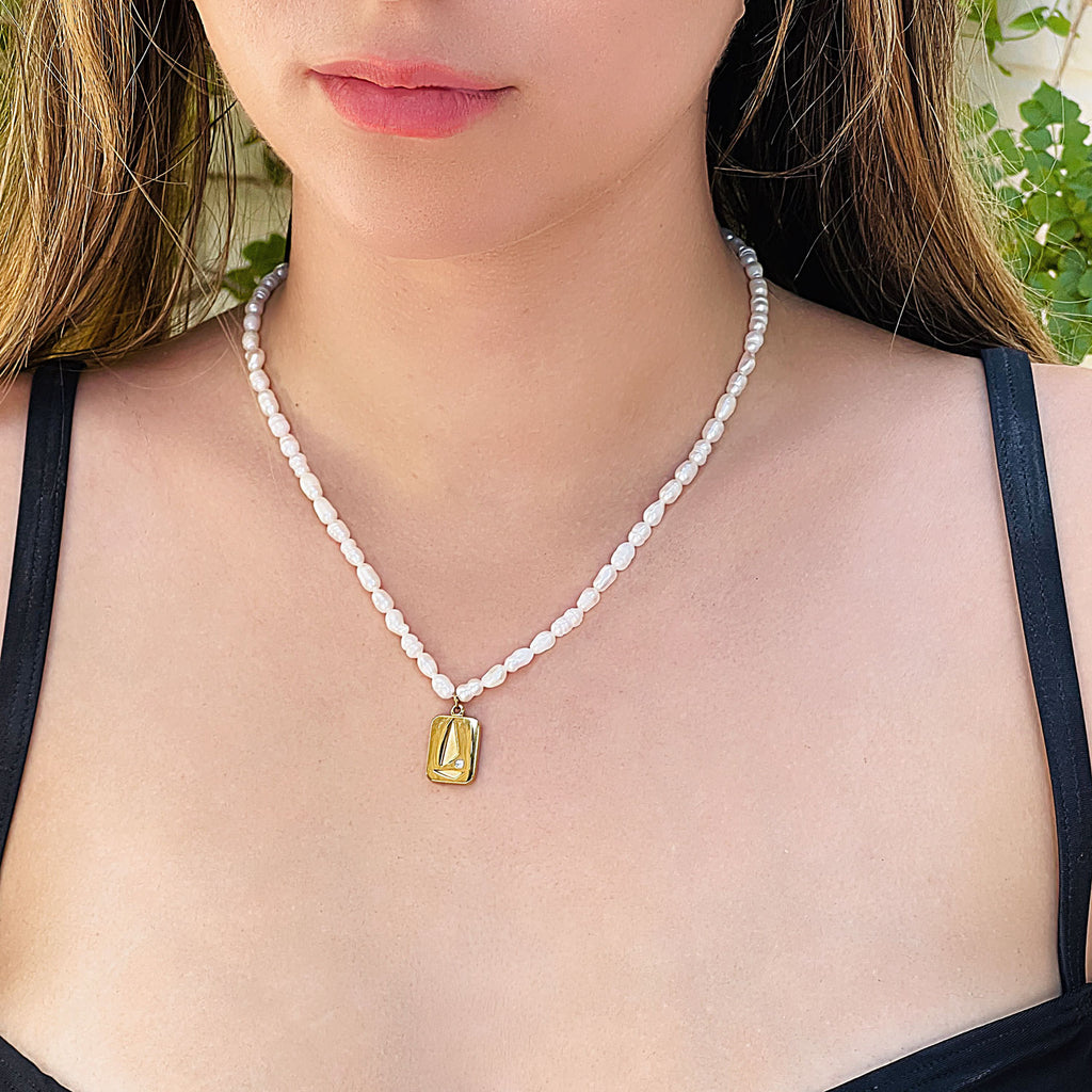 Girl wearing freshwater pearl beaded necklace with 18k gold plated pendant. Pendant is rectangular with abstract sailboat design with small cubic zirconia stone.