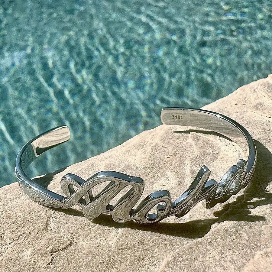 stainless steel bangle (silver color), cuff style bracelet carved script Aloha with engraved design motif, Sitting on ledge by pool.