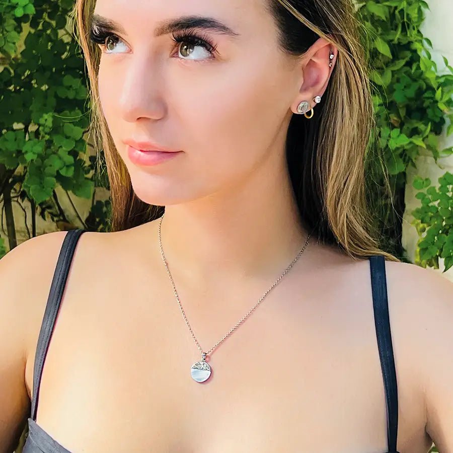 Girl wearing silver color stainless steel pendant necklace, pendant is mixed media circle, top half is an engraved patterned silver and bottom half is a semi-transparent light blue natural shell stone.
