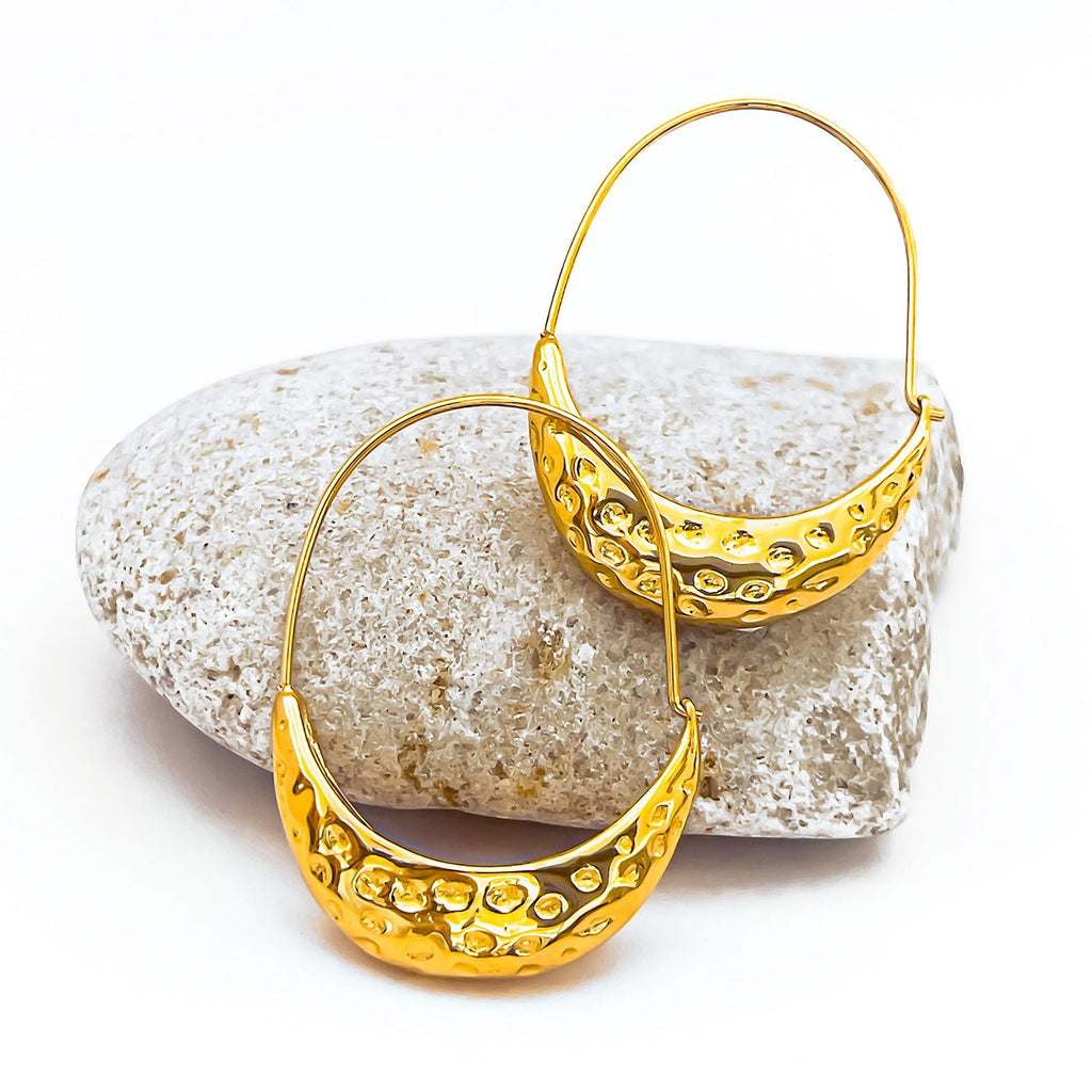 18k gold plated hammered hoop earrings. Thin hoop on top and thicker canoe-shaped and textured bottom half, displayed on a rock.